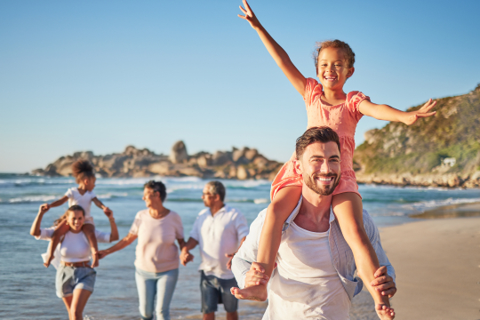 Vorne ein Vater mit einem M&auml;dchen auf den Schultern, links daneben eine Mutter mit einem weiteren Kind, und Oma und Opa am Strand laufend